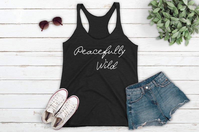 May include: A black tank top with the text "Peacefully Wild" in white script on the front. The tank top is laying on a white wooden surface with a pair of sunglasses, two white sneakers, and a pair of blue denim shorts.