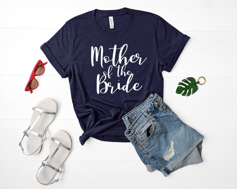 Puede incluir: Una camiseta azul marino con texto blanco que dice "Madre de la novia". La camiseta est&aacute; colocada sobre una superficie blanca con unos pantalones cortos de mezclilla, un par de sandalias plateadas y un llavero de hoja verde.