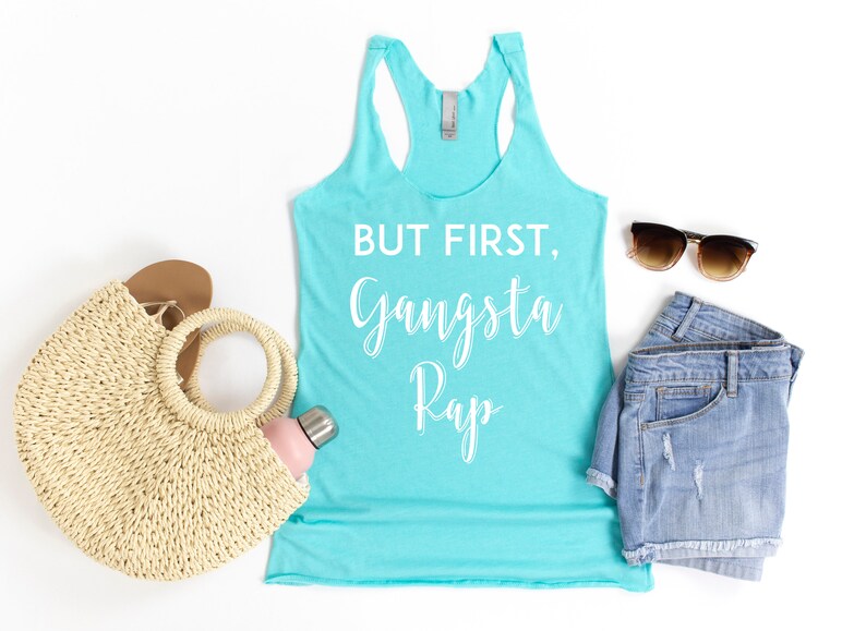 May include: A turquoise tank top with the text "But First, Gangsta Rap" in white lettering. The tank top is styled with denim shorts, sunglasses, a straw bag, and a water bottle.