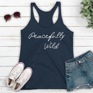 May include: A navy blue tank top with the text "Peacefully Wild" in white script on the front. The tank top is laying flat on a white wooden surface with a pair of white sneakers, a pair of sunglasses, and a pair of denim shorts.