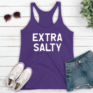 May include: Purple tank top with the text "EXTRA SALTY" in white lettering. The tank top is laying on a white wooden surface with a pair of white sneakers, a pair of sunglasses, and a pair of denim shorts.