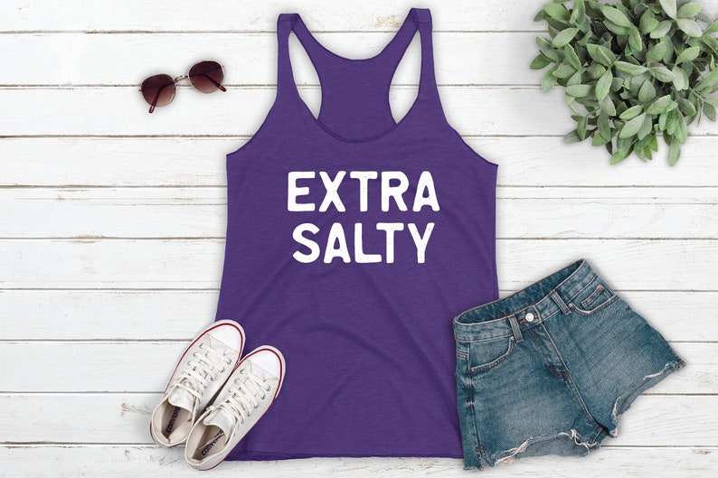 May include: Purple tank top with the text "EXTRA SALTY" in white lettering. The tank top is laying on a white wooden surface with a pair of white sneakers, a pair of sunglasses, and a pair of denim shorts.
