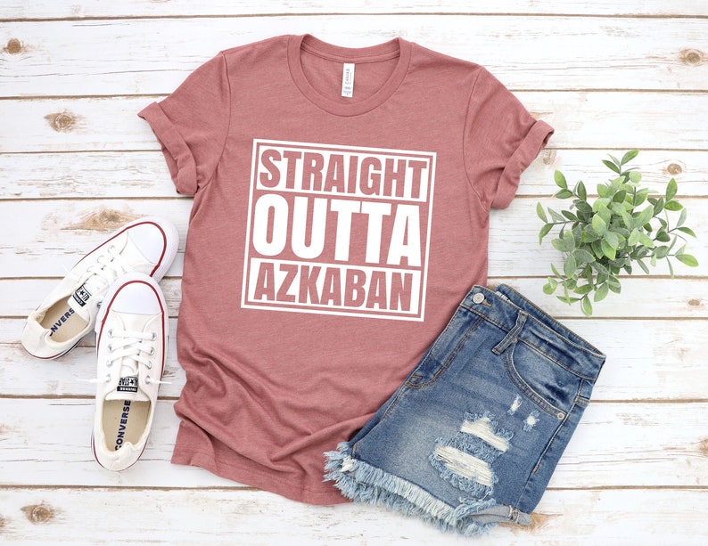 May include: A mauve t-shirt with the text "STRAIGHT OUTTA AZKABAN" printed in white on the front. The shirt is laid flat on a white wooden surface with a pair of white sneakers, a pair of denim shorts, and a small potted plant.