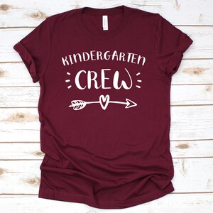 May include: Burgundy t-shirt with white text that reads "Kindergarten Crew" with a small heart and arrow graphic.