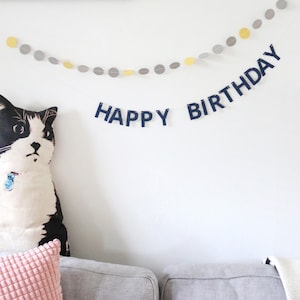 May include: A black and white cat-shaped pillow sits on a gray couch, with a pink and yellow pillow in front. A banner with the words "HAPPY BIRTHDAY" hangs on the white wall, along with a garland of yellow and gray circles.
