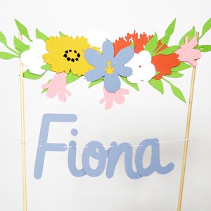 Puede incluir: Adorno de pastel floral colorido con el nombre "Fiona" en escritura azul claro. El adorno presenta una guirnalda de flores de papel en amarillo, azul, rojo, blanco y rosa, con hojas verdes. Montado sobre palitos de madera.