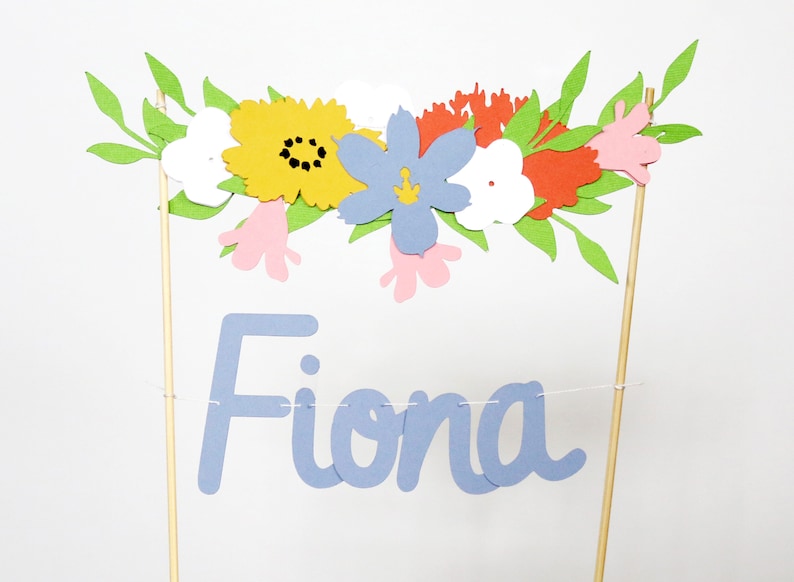 Puede incluir: Adorno de pastel floral colorido con el nombre "Fiona" en escritura azul claro. El adorno presenta una guirnalda de flores de papel en amarillo, azul, rojo, blanco y rosa, con hojas verdes. Montado sobre palitos de madera.