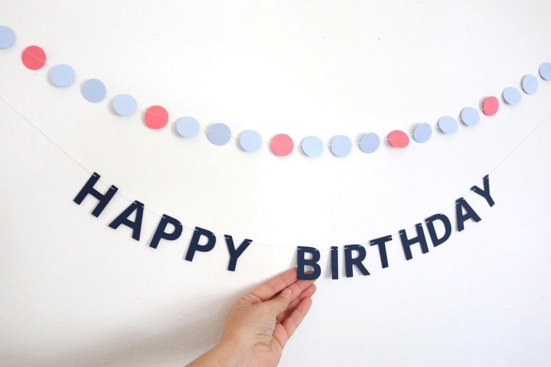 May include: A festive birthday banner with the words "HAPPY BIRTHDAY" in navy blue letters. Above the banner is a string of light blue and pink circular decorations, creating a celebratory atmosphere.
