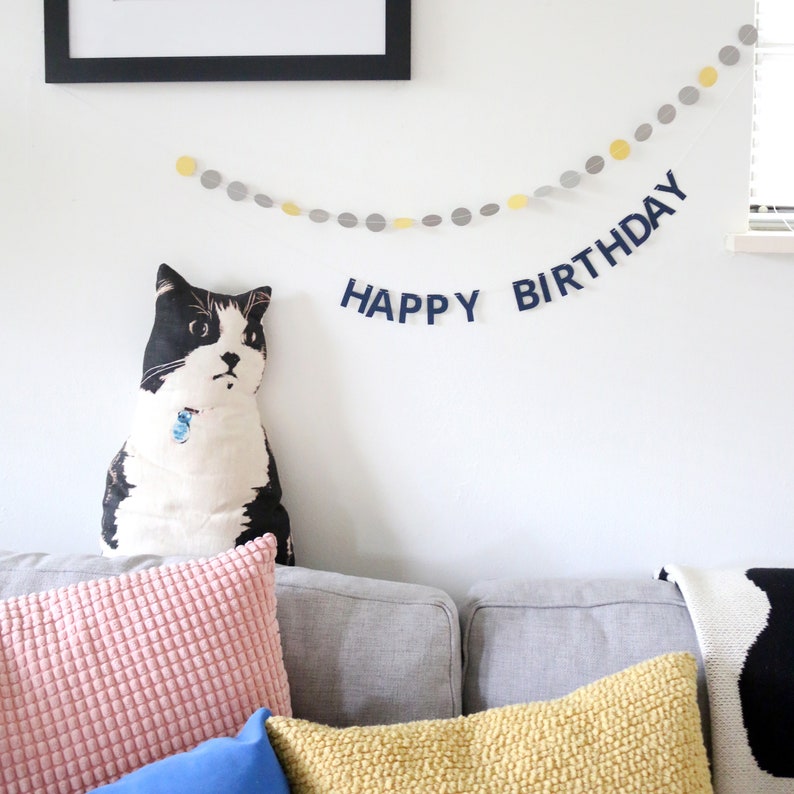 May include: A black and white cat-shaped pillow sits on a gray couch, with a pink and yellow pillow in front. A banner with the words "HAPPY BIRTHDAY" hangs on the white wall, along with a garland of yellow and gray circles.
