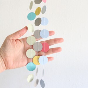 May include: A colorful garland of paper circles in various pastel shades, including pink, blue, yellow, and gray. The circles are strung together with white thread, creating a decorative hanging ornament. The garland is shown against a white background.