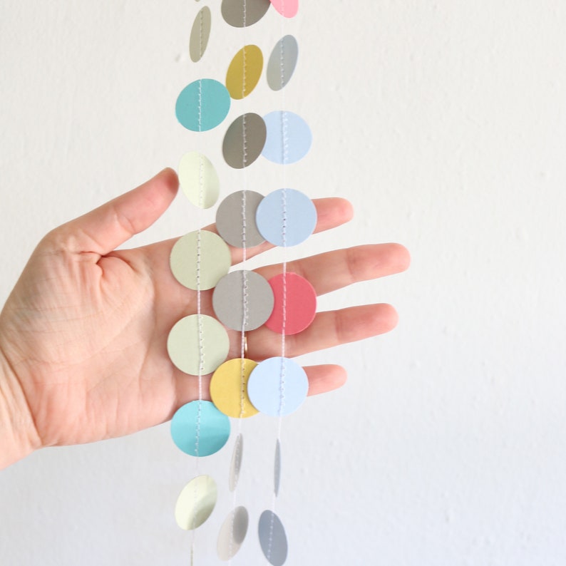 May include: A colorful garland of paper circles in various pastel shades, including pink, blue, yellow, and gray. The circles are strung together with white thread, creating a decorative hanging ornament. The garland is shown against a white background.