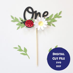 Puede incluir: Adorno para tarta con la palabra "One" en escritura negra, una mariquita roja, una margarita blanca y hojas verdes. El adorno está sobre un palito de madera. Un círculo azul con texto blanco dice "DIGITAL CUT FILE SVG DXXF".