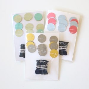 May include: Three packages of colorful paper garlands. Each garland features round paper cutouts in various pastel colors, strung together with white thread. Bundles of dark blue paper fringe are included.