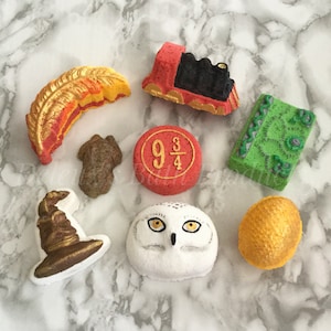 May include: A set of nine Harry Potter themed bath bombs. The bath bombs are shaped like a Hogwarts Express train, a Sorting Hat, a golden snitch, a snowy owl, a feather, a book, and a platform 9 3/4 sign.