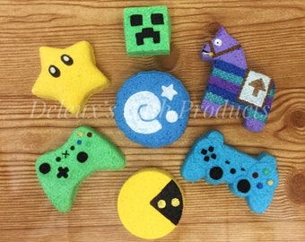 May include: A set of six colorful bath bombs shaped like popular video game characters. The bath bombs are green, yellow, blue, purple, and black. The characters include a Minecraft Creeper, a Pac-Man ghost, a Super Mario star, a Fortnite Llama, and two video game controllers.