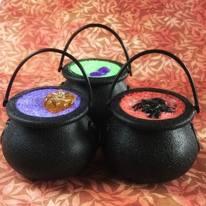 Peut inclure: Trois récipients noirs en forme de chaudron avec des poignées. Chaque chaudron est rempli de sable coloré et d'un bonbon sur le thème d'Halloween. Les chaudrons sont sur une surface orange et brune à motifs.