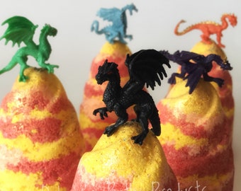May include: Close-up of several colorful bath bombs, each topped with a small plastic dragon figurine. The bath bombs are yellow, red, and orange, with a glittery texture. The dragons are green, blue, orange, purple, and black.