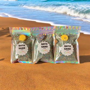 May include: Three iridescent zip-lock bags filled with colourful bath sand, each sealed with a yellow flower-shaped sticker. Each bag has a white label with the words "BATH SAND" and "100 g". The bags are on a sandy beach with the ocean in the background.