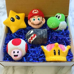 May include: A set of six bath bombs shaped like characters from the Super Mario Bros. video game. The bath bombs are yellow, red, green, black, white, and pink. They are in a white box with blue shredded paper.