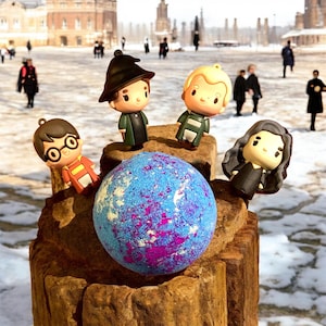 May include: A blue and purple bath bomb is displayed on a wooden plinth, with character figurines around it. The figurines are styled after characters from the Harry Potter series. The background shows a snowy outdoor scene.