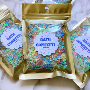 May include: Three gold foil pouches filled with colorful bath confetti. Each pouch has a white label with the text "Bath Confetti 100 grams" in blue and purple text.