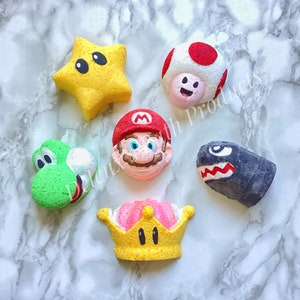May include: Six colorful bath bombs shaped like characters from the Super Mario Bros. video game franchise. The bath bombs are yellow, red, green, black, pink, and white.