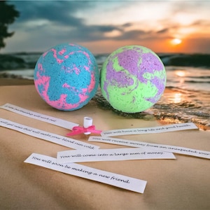 May include: Two colorful bath bombs, one blue and pink, the other green and purple, sit on a sandy beach. Fortune slips with printed messages are scattered around them. A pink ribbon and a small white tube are also present. The background shows the ocean and a sunset.