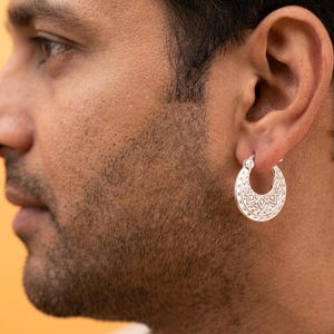 Peut inclure: Une cr&eacute;ole en argent orn&eacute;e d'un motif g&eacute;om&eacute;trique complexe et d&eacute;taill&eacute; grav&eacute; sur toute sa surface. La boucle d'oreille est de forme circulaire avec une finition m&eacute;tallique polie, pr&eacute;sentant un design sym&eacute;trique r&eacute;p&eacute;t&eacute; sur la face externe.