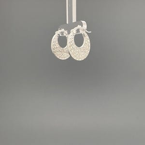 Peut inclure: Une paire de cr&eacute;oles en argent pr&eacute;sentant un motif g&eacute;om&eacute;trique textur&eacute; sur la surface ext&eacute;rieure. Les boucles d'oreilles sont dot&eacute;es d'un m&eacute;canisme de fermeture &agrave; charni&egrave;re sur le dessus et d'un design circulaire qui s'affine vers le bas.