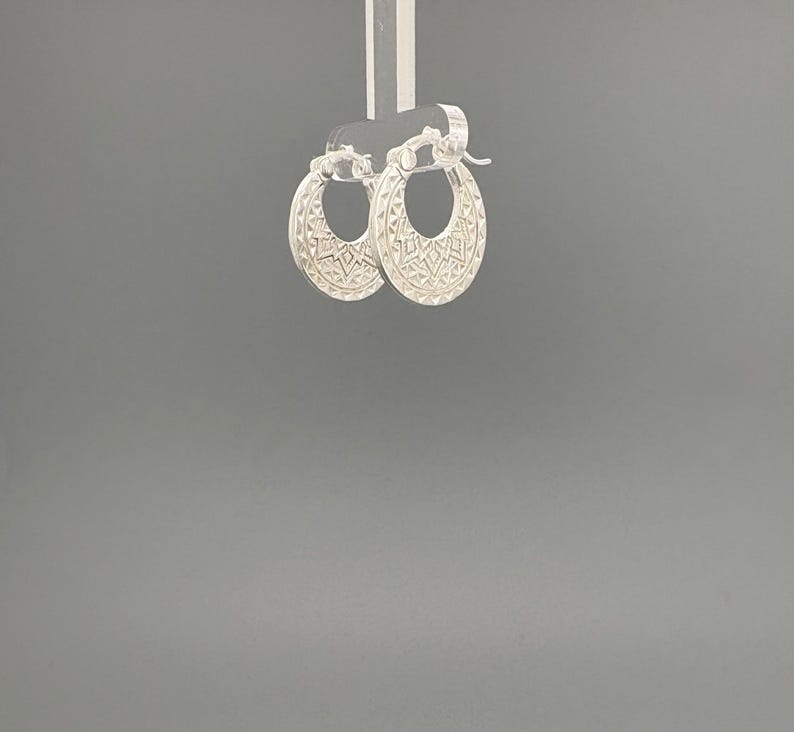 Peut inclure: Une paire de cr&eacute;oles en argent pr&eacute;sentant un motif g&eacute;om&eacute;trique textur&eacute; sur la surface ext&eacute;rieure. Les boucles d'oreilles sont dot&eacute;es d'un m&eacute;canisme de fermeture &agrave; charni&egrave;re sur le dessus et d'un design circulaire qui s'affine vers le bas.