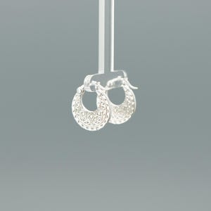 Peut inclure: Une paire de petites cr&eacute;oles en argent pr&eacute;sentant un design circulaire avec des d&eacute;coupes g&eacute;om&eacute;triques complexes et des motifs grav&eacute;s sur le bord ext&eacute;rieur. Les boucles d'oreilles sont suspendues &agrave; un pr&eacute;sentoir en acrylique transparent.