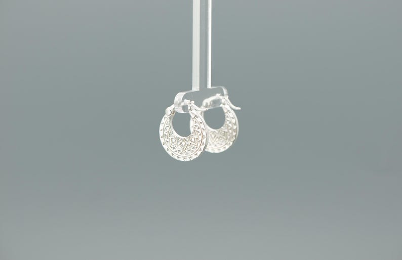Peut inclure: Une paire de petites cr&eacute;oles en argent pr&eacute;sentant un design circulaire avec des d&eacute;coupes g&eacute;om&eacute;triques complexes et des motifs grav&eacute;s sur le bord ext&eacute;rieur. Les boucles d'oreilles sont suspendues &agrave; un pr&eacute;sentoir en acrylique transparent.