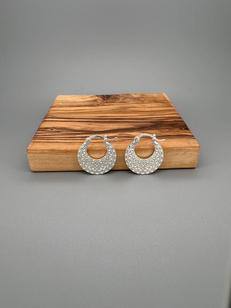 Peut inclure: Une paire de cr&eacute;oles en argent pr&eacute;sentant un motif g&eacute;om&eacute;trique textur&eacute; sur la surface avant. Les boucles d'oreilles ont une forme circulaire avec un m&eacute;canisme de fermeture &agrave; charni&egrave;re, arborant une finition m&eacute;tallique et des d&eacute;tails triangulaires.
