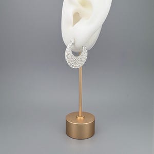 Peut inclure: Une boucle d'oreille cr&eacute;ole en argent en forme de croissant avec un motif g&eacute;om&eacute;trique textur&eacute; compos&eacute; de petits triangles et de lignes. La boucle est pr&eacute;sent&eacute;e sur un support blanc en forme d'oreille fix&eacute; sur une base cylindrique dor&eacute;e.
