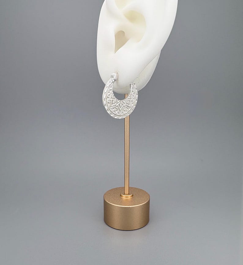 Peut inclure: Une boucle d'oreille cr&eacute;ole en argent en forme de croissant avec un motif g&eacute;om&eacute;trique textur&eacute; compos&eacute; de petits triangles et de lignes. La boucle est pr&eacute;sent&eacute;e sur un support blanc en forme d'oreille fix&eacute; sur une base cylindrique dor&eacute;e.