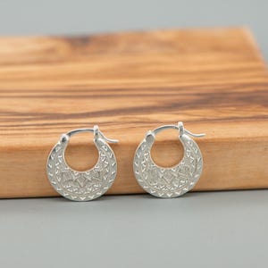 Peut inclure: Une paire de cr&eacute;oles en argent pr&eacute;sentant une surface textur&eacute;e avec des motifs g&eacute;om&eacute;triques complexes. Les boucles d'oreilles ont une forme circulaire avec une fermeture &agrave; tige articul&eacute;e, arborant une finition m&eacute;tallique et des gravures d&eacute;taill&eacute;es.