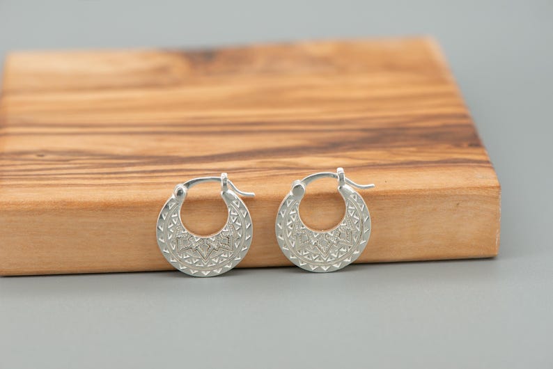 Peut inclure: Une paire de cr&eacute;oles en argent pr&eacute;sentant une surface textur&eacute;e avec des motifs g&eacute;om&eacute;triques complexes. Les boucles d'oreilles ont une forme circulaire avec une fermeture &agrave; tige articul&eacute;e, arborant une finition m&eacute;tallique et des gravures d&eacute;taill&eacute;es.