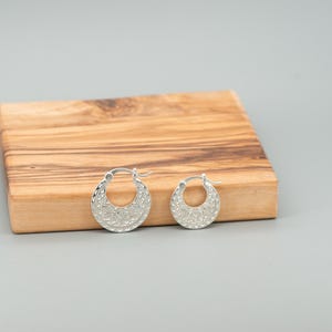 Peut inclure: Une paire de cr&eacute;oles en argent pr&eacute;sentant un design textur&eacute; en forme de croissant avec des motifs complexes. Les boucles d'oreilles sont circulaires avec un m&eacute;canisme de fermeture &agrave; charni&egrave;re, affichant une finition m&eacute;tallique polie.