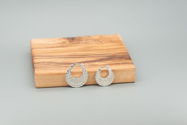 Peut inclure: Une paire de cr&eacute;oles en argent pr&eacute;sentant un design textur&eacute; en forme de croissant avec des motifs complexes. Les boucles d'oreilles sont circulaires avec un m&eacute;canisme de fermeture &agrave; charni&egrave;re, affichant une finition m&eacute;tallique polie.
