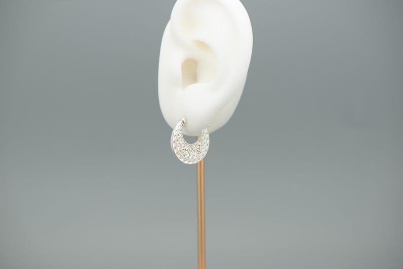 Peut inclure: Une petite boucle d'oreille cr&eacute;ole argent&eacute;e pr&eacute;sentant un design en forme de croissant avec un motif filigrane en relief complexe sur toute sa surface. La boucle d'oreille est pr&eacute;sent&eacute;e sur un support en forme d'oreille blanche.