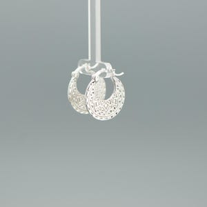 Peut inclure: Une paire de cr&eacute;oles en argent pr&eacute;sentant un motif d&eacute;coup&eacute; d&eacute;taill&eacute; et complexe sur toute la surface. Les boucles d'oreilles ont une forme circulaire avec un m&eacute;canisme de fermeture &agrave; charni&egrave;re sur le dessus, arborant une finition m&eacute;tallique.