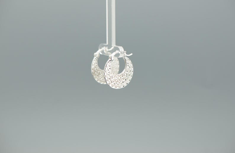 Peut inclure: Une paire de cr&eacute;oles en argent pr&eacute;sentant un motif d&eacute;coup&eacute; d&eacute;taill&eacute; et complexe sur toute la surface. Les boucles d'oreilles ont une forme circulaire avec un m&eacute;canisme de fermeture &agrave; charni&egrave;re sur le dessus, arborant une finition m&eacute;tallique.