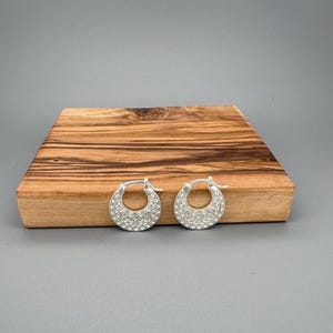 Peut inclure: Une paire de cr&eacute;oles en argent pr&eacute;sentant une surface textur&eacute;e avec un motif g&eacute;om&eacute;trique r&eacute;p&eacute;titif compos&eacute; de petits triangles et de losanges. Les boucles d'oreilles sont dot&eacute;es d'un fermoir &agrave; charni&egrave;re et d'une finition mate.