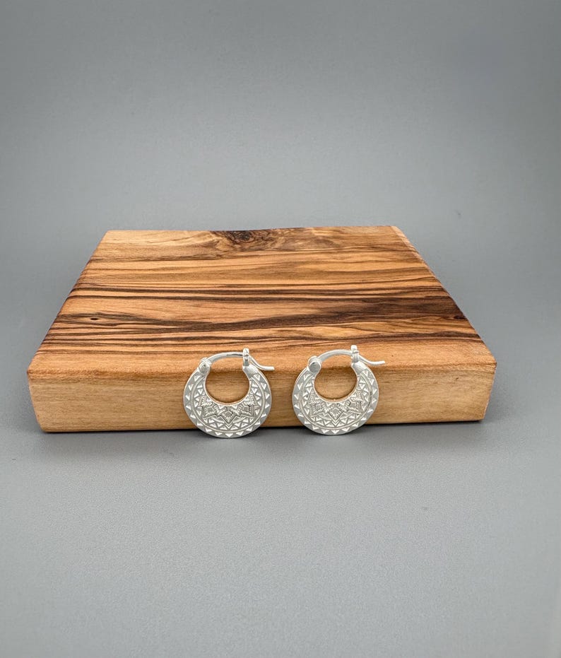 Peut inclure: Une paire de cr&eacute;oles en argent pr&eacute;sentant une surface textur&eacute;e avec un motif g&eacute;om&eacute;trique r&eacute;p&eacute;titif compos&eacute; de petits triangles et de losanges. Les boucles d'oreilles sont dot&eacute;es d'un fermoir &agrave; charni&egrave;re et d'une finition mate.