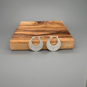 Peut inclure: Une paire de cr&eacute;oles en argent dot&eacute;es d'une section inf&eacute;rieure large en forme de croissant. La surface de chaque boucle d'oreille est orn&eacute;e d'un motif g&eacute;om&eacute;trique r&eacute;p&eacute;titif compos&eacute; de petits triangles et de lignes, avec un fermoir &agrave; charni&egrave;re.