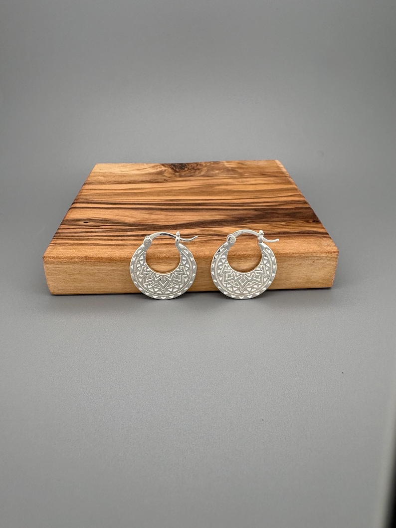 Peut inclure: Une paire de cr&eacute;oles en argent dot&eacute;es d'une section inf&eacute;rieure large en forme de croissant. La surface de chaque boucle d'oreille est orn&eacute;e d'un motif g&eacute;om&eacute;trique r&eacute;p&eacute;titif compos&eacute; de petits triangles et de lignes, avec un fermoir &agrave; charni&egrave;re.