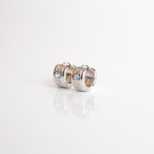 Peut inclure: Une paire de boucles d'oreilles créoles en argent avec un motif décoratif et une pierre précieuse bleu clair sur chaque boucle d'oreille.