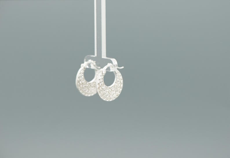 Peut inclure: Une paire de petites cr&eacute;oles en argent pr&eacute;sentant un motif g&eacute;om&eacute;trique textur&eacute; et d&eacute;taill&eacute; sur la surface ext&eacute;rieure. Les boucles d'oreilles sont de forme circulaire avec un m&eacute;canisme de fermeture &agrave; charni&egrave;re et une finition m&eacute;tallique brillante.
