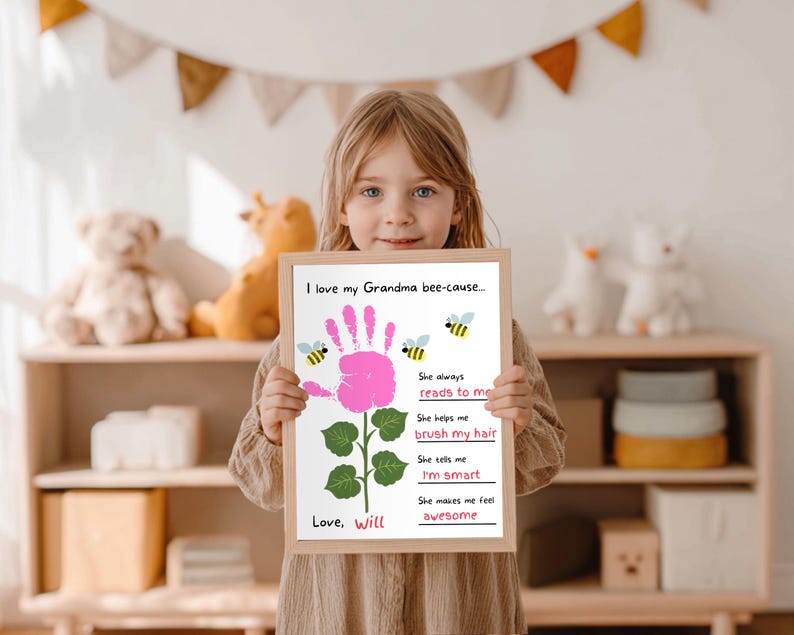 May include: A child holds a framed print with the text "I love my Grandma bee-cause..." and a pink handprint flower design. The print includes the phrases "She always reads to me," "brush my hair," "I'm smart," and "awesome."