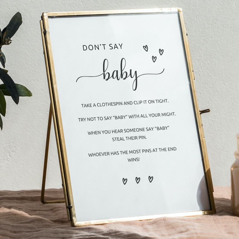 Może przedstawiać: Złota ramka z napisem "DON'T SAY baby" czarnym skryptem. Znak zawiera instrukcje do gry na baby shower, z ilustracjami serc. Rama wykonana jest ze złocistego metalu.